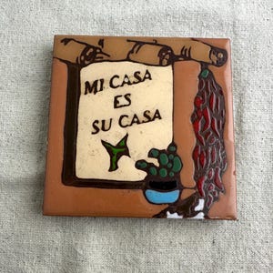 May include: Square ceramic tile with a brown border and a cream-colored center. The center features the text "MI CASA ES SU CASA" in black, with a green plant and a blue pot. Decorative tile.