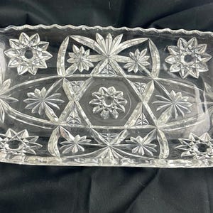May include: A rectangular clear glass serving tray featuring a pressed pattern of multiple starburst motifs, geometric triangles, and a scalloped edge. The design includes a central starburst surrounded by radiating lines and smaller starbursts.