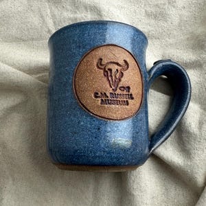 May include: Blue ceramic mug with a brown circular emblem featuring a bull skull and the text "C.M. Russell Museum." The mug has a curved handle and a speckled finish. The mug is on a beige cloth.