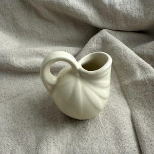 May include: Off-white ceramic creamer with a curved handle and a rounded body. The creamer has a fluted design on the exterior and a small opening for pouring. The handle is a loop shape.