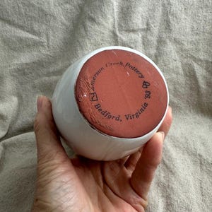Puede incluir: Un jarr&oacute;n de cer&aacute;mica blanco con forma redondeada. La base es de color marr&oacute;n rojizo y presenta el texto "Emerson Creek Pottery '93 Bedford, Virginia" en una disposici&oacute;n circular. El jarr&oacute;n est&aacute; sostenido en una mano.