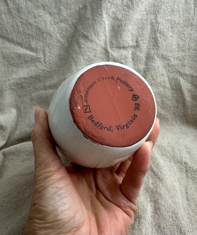 Puede incluir: Un jarr&oacute;n de cer&aacute;mica blanco con forma redondeada. La base es de color marr&oacute;n rojizo y presenta el texto "Emerson Creek Pottery '93 Bedford, Virginia" en una disposici&oacute;n circular. El jarr&oacute;n est&aacute; sostenido en una mano.