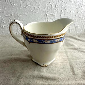 May include: Cream-colored ceramic creamer with a blue and gold decorative band. The creamer has a curved handle and a spout for pouring. The design includes floral and leaf patterns, adding a touch of elegance to the piece.