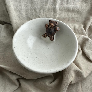 May include: A round, off-white ceramic bowl with a speckled finish and a small, dark brown, three-dimensional bear figure attached to the inner rim. The bowl has a smooth, curved interior surface and a simple, circular design.