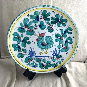 May include: A decorative ceramic plate featuring a hand-painted rooster design. The plate has a white base with a teal rooster, teal and red floral accents, and a yellow border. The rim has a textured, rope-like design.