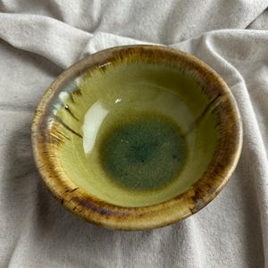 May include: A small, handmade ceramic bowl with a glossy finish. The bowl features a gradient of colors, with a dark green center that fades to a light green and then to a brown rim. The bowl is approximately 4 inches in diameter.
