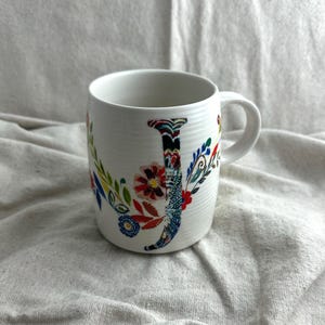 Op de afbeelding: Witte keramische mok met de letter "J", versierd met een kleurrijk bloemmotief. De mok is versierd met bloemen en bladeren in verschillende kleuren. Hij heeft een gebogen handvat en is geschikt voor warme dranken.