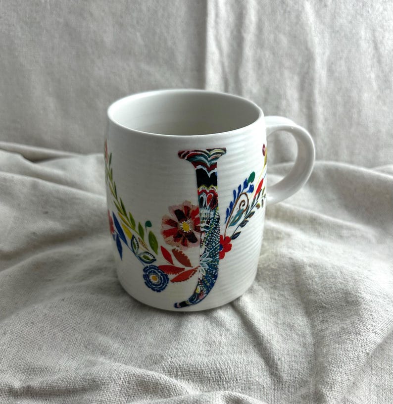 Op de afbeelding: Witte keramische mok met de letter "J", versierd met een kleurrijk bloemmotief. De mok is versierd met bloemen en bladeren in verschillende kleuren. Hij heeft een gebogen handvat en is geschikt voor warme dranken.