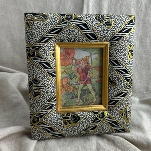May include: A rectangular photo frame with a gold-colored inner frame. The outer frame is covered in a black, white, and gold patterned fabric. The picture inside the frame depicts a person with a bow and arrow.