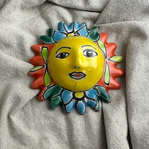 May include: A ceramic sun face wall hanging. The sun has a yellow face with black eyes and a red mouth. The sun's rays are red, with blue and green accents. The sun is a decorative item.