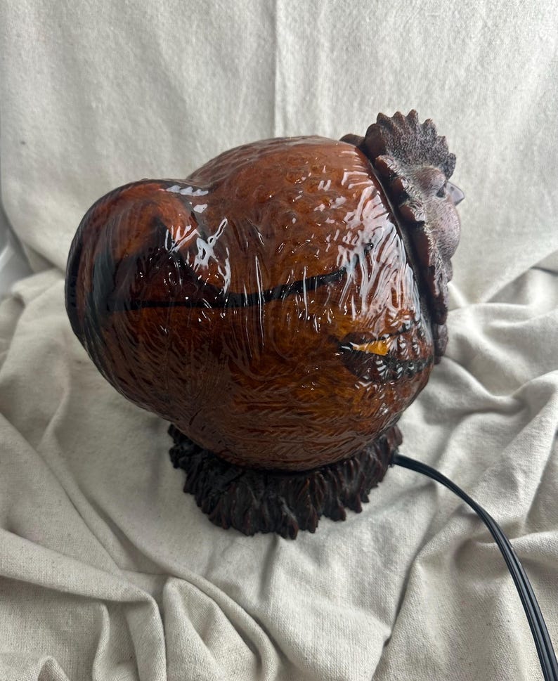 May include: A brown rooster-shaped lamp with a glossy finish. The lamp features a detailed textured design, including the rooster's comb and feathers. The lamp sits on a dark brown base and has a black power cord.