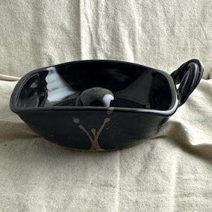 May include: A black ceramic bowl with a glossy finish and a handle. The bowl has a rounded shape with a small, round object in the center. It features a decorative, abstract design in a light brown color.