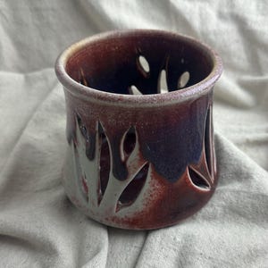May include: A handmade ceramic candle holder with a unique leaf-shaped cut-out design. The candle holder features a gradient of colors, transitioning from a light grey at the base to deep burgundy and purple at the rim. The interior is dark.