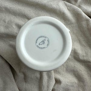 May include: A white ceramic mug base with a circular stamp featuring a bird illustration and text. The stamp reads "MICROWAVE & DISHWASHER SAFE" and "FABRIQUE EN CHINE". The mug is set against a neutral fabric background.