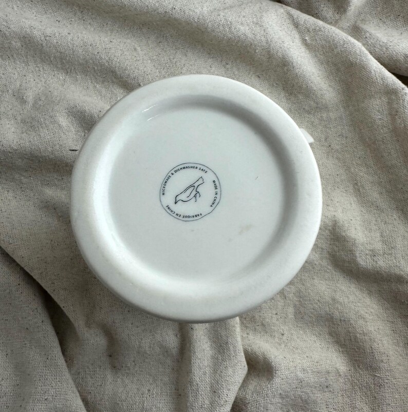 May include: A white ceramic mug base with a circular stamp featuring a bird illustration and text. The stamp reads "MICROWAVE & DISHWASHER SAFE" and "FABRIQUE EN CHINE". The mug is set against a neutral fabric background.
