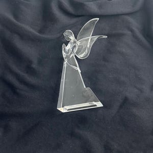May include: A clear, crystal angel figurine with outstretched wings. The angel is standing on a triangular base. The wings and body are transparent, allowing light to pass through. The figurine is set against a dark background.