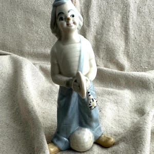 May include: A ceramic clown figurine wearing a light blue hat and overalls, holding a small hat and a spotted bag. The clown has a white striped shirt and stands next to a white ball. The figurine has a smiling face.
