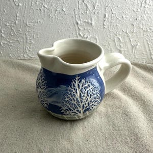 May include: A ceramic pitcher with a white interior and handle. The exterior features a blue band with a white tree design. The pitcher has a spout and is suitable for serving beverages. The overall design is rustic and handmade.