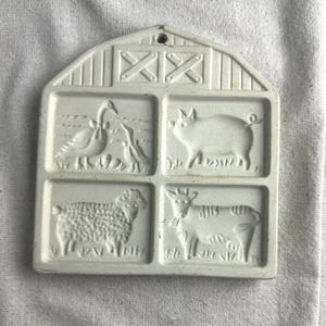 May include: White ceramic cookie mold shaped like a barn with four squares. Each square has a different farm animal design: a goose, a pig, a sheep, and a cow.