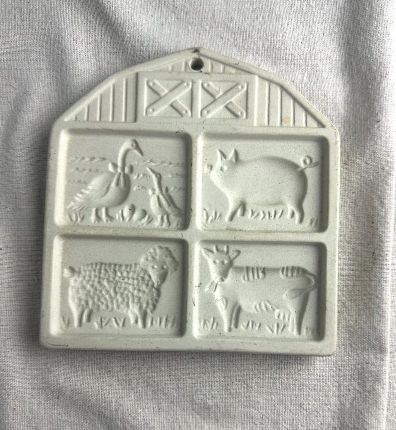 May include: White ceramic cookie mold shaped like a barn with four squares. Each square has a different farm animal design: a goose, a pig, a sheep, and a cow.