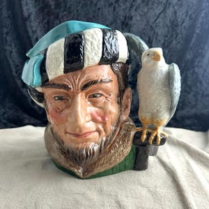 May include: A ceramic figurine of a man with a blue and white hat and a beard, holding a falcon on his shoulder. The figurine is painted in realistic detail and has a green base.