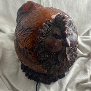 May include: A brown, carved chicken figurine with a detailed comb, wattle, and feather texture. The sculpture has a glossy finish and a dark brown base. A black cord extends from the base.