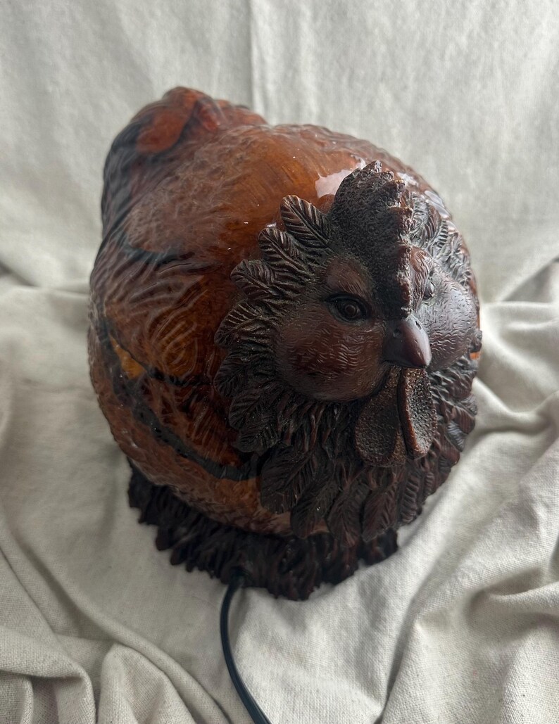 May include: A brown, carved chicken figurine with a detailed comb, wattle, and feather texture. The sculpture has a glossy finish and a dark brown base. A black cord extends from the base.