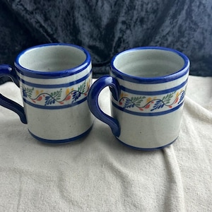 May include: Two white ceramic mugs with blue rims and a floral design in blue, red, and yellow. The mugs have blue handles.