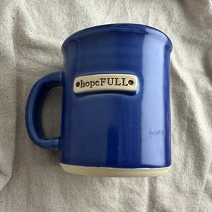 May include: A deep blue ceramic mug with a curved handle. The mug has the word "hopeFULL" on a small, rectangular plaque. The base of the mug is a light beige colour, and it sits against a neutral background.
