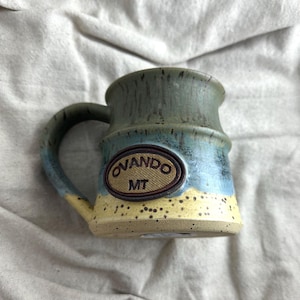 May include: A handcrafted ceramic mug with a distinctive design. The mug has a green top, blue middle, and yellow base. An oval emblem with "Ovando MT" is on the front. The handle and interior are green. The mug is on a neutral fabric background.