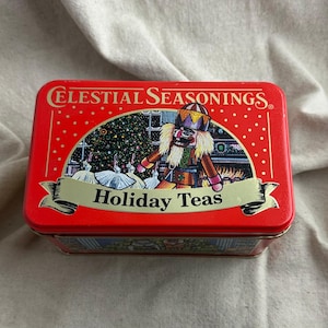 May include: A rectangular red metal tin for Celestial Seasonings Holiday Teas. The lid features a festive illustration of a nutcracker and ballerinas in front of a fireplace and Christmas tree, with the text Celestial Seasonings Holiday Teas.