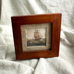 May include: A square wooden frame with a medium brown finish containing a small print of a sailing ship on the ocean. The print is mounted on a textured, light tan hessian fabric background, creating a layered appearance within the frame.
