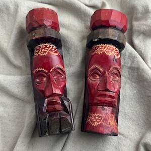 May include: Two carved wooden sculptures with red faces and geometric details. The sculptures have a dark brown base and a red top. The faces have carved features and are set against a neutral background.