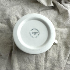May include: White ceramic mug with a handle, featuring a blue bird logo and text on the bottom. The mug is set against a neutral-colored fabric background. The text reads "MICROWAVE & DISHWASHER SAFE" and "MADE IN CHINA".