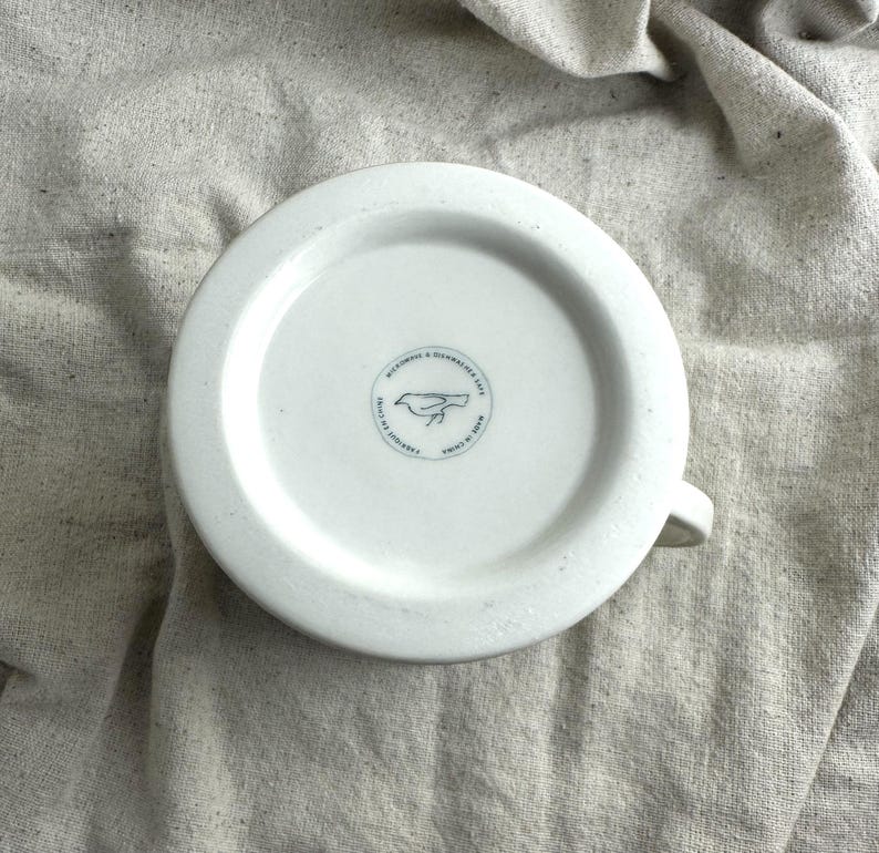 May include: White ceramic mug with a handle, featuring a blue bird logo and text on the bottom. The mug is set against a neutral-colored fabric background. The text reads "MICROWAVE & DISHWASHER SAFE" and "MADE IN CHINA".