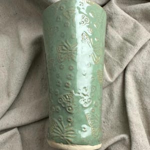 May include: A tall, light green ceramic tumbler with a textured surface. The tumbler is adorned with embossed butterfly and floral patterns. The base is a lighter, unglazed color. The tumbler is approximately 6 inches tall.