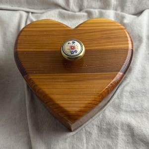 May include: A heart-shaped wooden box with a floral knob. The box is made of light brown wood with visible grain. The knob is gold-toned with a white center featuring pink, blue, and yellow flowers.