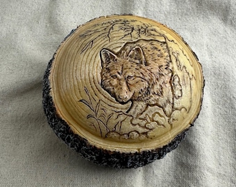 May include: A round, wooden carving featuring a detailed depiction of a wolf. The wolf is carved in relief, with intricate details in its fur and facial features. The wood has a natural, light brown color with a dark, textured bark edge.