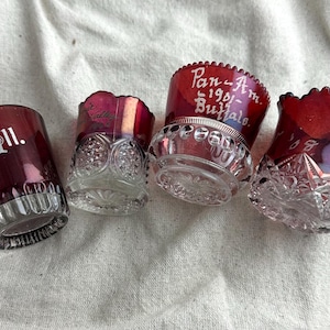 May include: Four antique ruby red glass tumblers with etched designs and text. One tumbler reads "Ill. 1906." Another reads "Pan-Am. 1901-Buffalo." The glasses have clear, cut-glass bases and decorative rims.