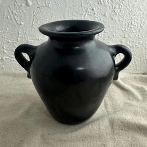 May include: A black ceramic vase with two handles and a wide opening. The vase has a smooth, glossy finish and a rounded body. The vase is sitting on a beige surface.