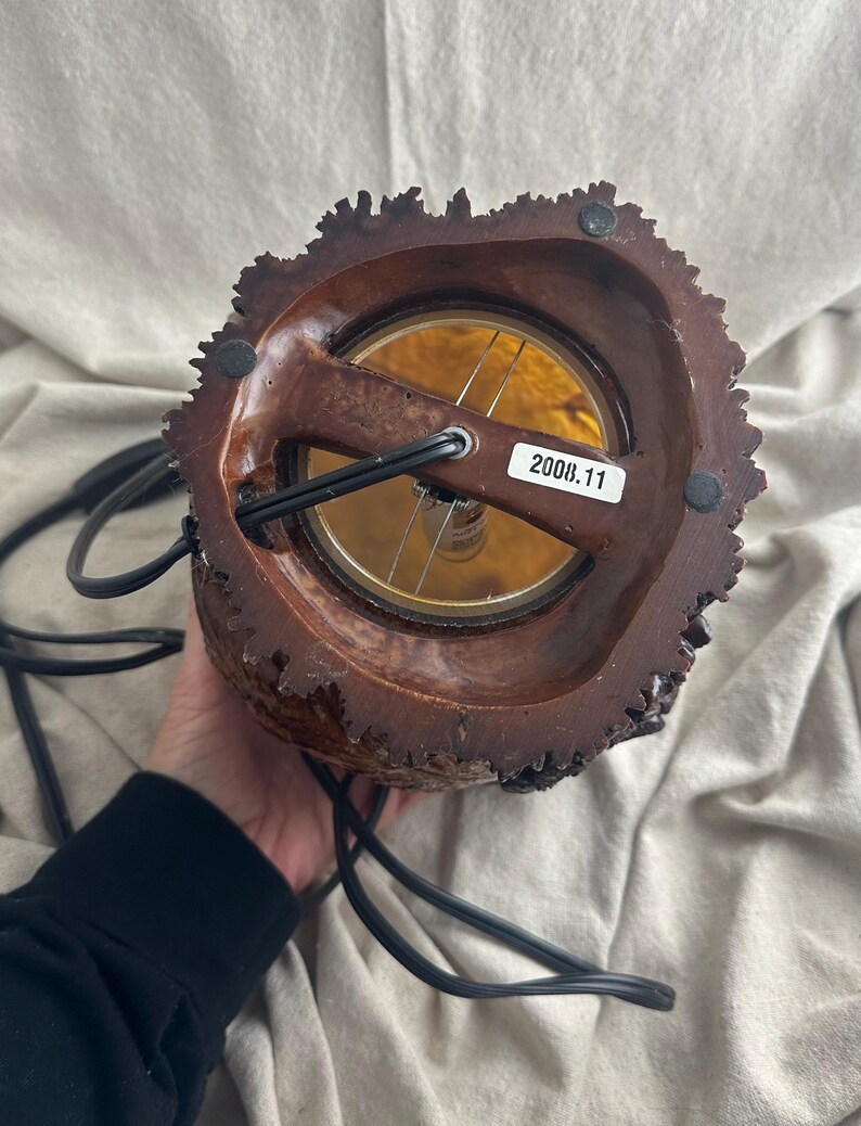 May include: A brown, rustic-style lamp with a textured, bark-like exterior. The lamp features a warm, amber-colored light source and a black power cord. The label on the lamp reads "2008.11".