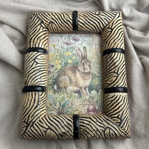 May include: Rectangular picture frame with a detailed zebra-stripe pattern in beige and black. The frame holds a watercolor image of a brown rabbit in a field of flowers. Black bands accent the frame's corners.