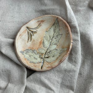 May include: A small, handmade ceramic dish with a leaf design. The dish is oval-shaped with a light beige background and a brown rim. A detailed leaf imprint is visible, with a smaller floral design on the left side.