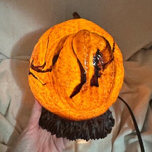 May include: A glowing orange pumpkin-shaped lamp with dark brown accents. The lamp has a textured surface and sits on a dark brown base. A black cord extends from the base. The lamp is lit from within.