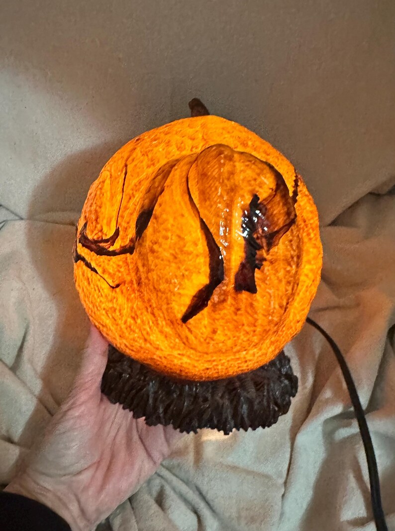 May include: A glowing orange pumpkin-shaped lamp with dark brown accents. The lamp has a textured surface and sits on a dark brown base. A black cord extends from the base. The lamp is lit from within.