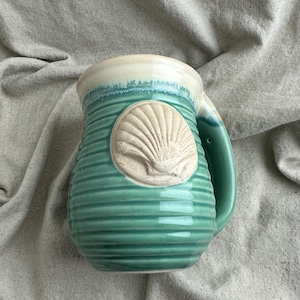 May include: A ceramic mug featuring a ribbed, teal-green glazed body with a smooth, cream-coloured rim. A raised, unglazed beige seashell relief is centred on the front. The mug includes a sturdy handle on the right side with a matching teal glaze.