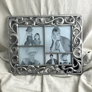 May include: Silver-colored multi-photo frame with an ornate, scrolling design. The frame holds five black and white photographs. The photos feature people of different ages. The frame is rectangular with a decorative border.