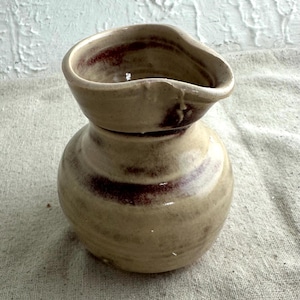 May include: A handmade ceramic vase with a bulbous base and a flared top. The vase is primarily beige with dark red-brown accents around the rim and base. The unique shape and color variations suggest a rustic, artisanal aesthetic.