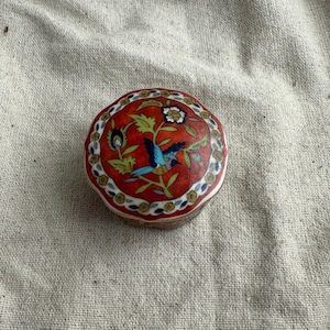 May include: A small, round, decorative box with a red background and a detailed bird and floral design. The bird is blue and the flowers are white and yellow. The box has a decorative border.