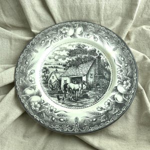 May include: A round ceramic plate featuring a black and white transferware scene of a blacksmith shop with a horse, a person, and a dog. The border is decorated with a detailed pattern of fruits, wheat stalks, and flowers in a monochromatic style.
